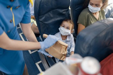 Koruyucu yüz maskesi takmış şirin bir kız uçakta küçük bir yolcuya yemek servisi yapan hostese bakıyor. Covid19 salgını sırasında uçakla seyahat etmek