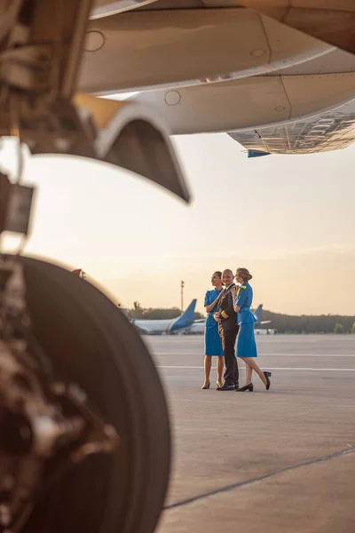 Full length shot of male pilot posing for photoshoot together with two air stewardesses in blue uniform, standing in an airport terminal at sunset — Stock Photo, Image