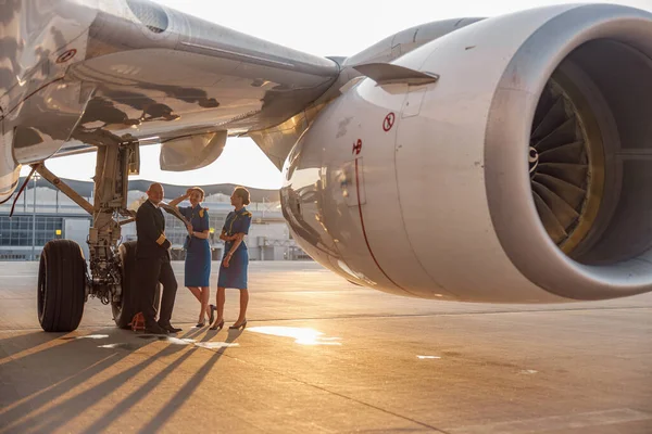 Happy pilot and two pretty stewardesses standing together, leaning on an airplane and smiling at camera after landing or before departure — Stock Photo, Image