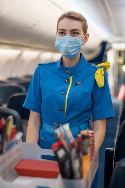 Letuška v ochranné masce se dívá stranou a podává jídlo cestujícím v letadle. Letuška chodící s vozíkem v uličce — Stock fotografie