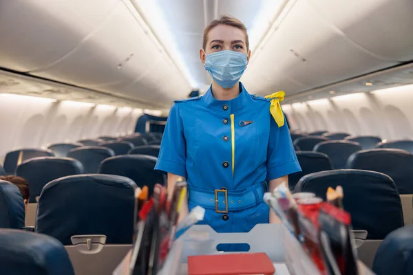 Azafata con mascarilla protectora y uniforme azul sirviendo comida a los pasajeros en los aviones. Azafata caminando con carro en el pasillo — Foto de Stock