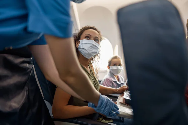 Žena v ochranné masce obličeje a její dcera při pohledu na ženské letušky pomáhá jim přizpůsobit a těsné bezpečnostní pásy v letadle pro bezpečný výlet — Stock fotografie