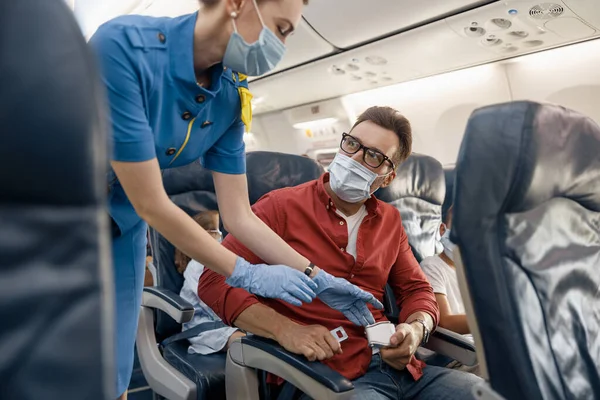 女性の客室乗務員を見て保護面マスクの男は、彼が安全旅行のために飛行機のシートベルトを調整し、タイトアップするのを助けます — ストック写真