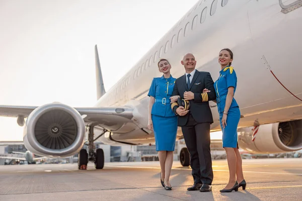 Full length shot of excited male pilot posing together with two air hostesses in blue uniform in front of an airplane in terminal at sunset