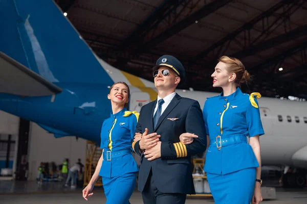 Happy pilot in uniform and aviator sunglasses walking together with two air stewardesses in blue uniform in front of big passenger airplane in airport hangar