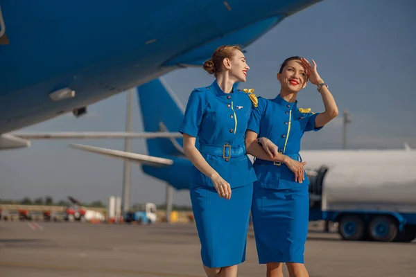 Dvě veselé letušky v jasně modré uniformě kráčející venku před osobními letadly za slunečného dne — Stock fotografie