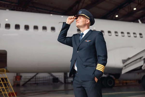 Inspired pilot in uniform looking away, adjusting his hat, standing in front of big passenger airplane in airport hangar