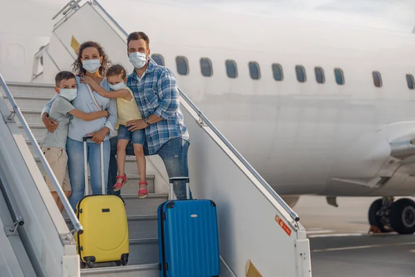 Family in protective masks, parents with two little children standing on airstair, boarding the plane during Covid19 pandemic