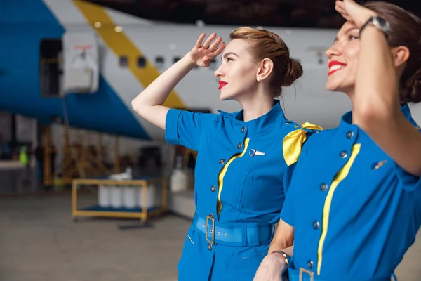 Portrét dvou krásných letušek ve stylové modré uniformě, usmívajících se při pohledu na oblohu, stojících společně před osobními letadly v hangáru na letišti — Stock fotografie