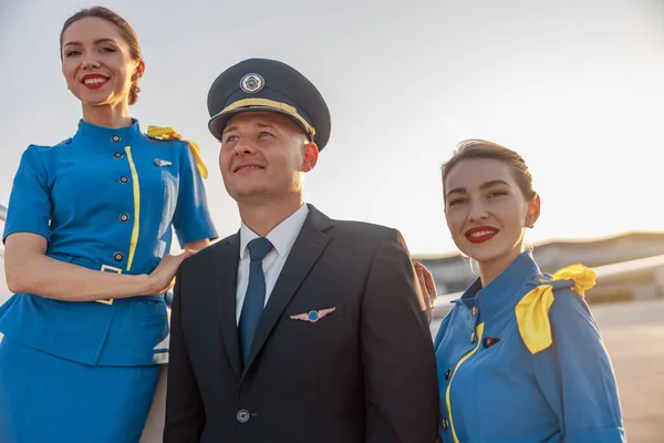Portrait of male pilot wearing hat posing together with two air hostesses in blue uniform outdoors