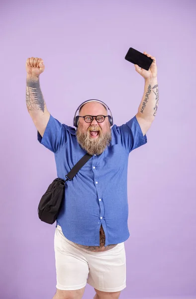 Opgewonden dikke man met hoofdtelefoon heft handen en mobiele telefoon poseren op paarse achtergrond — Stockfoto