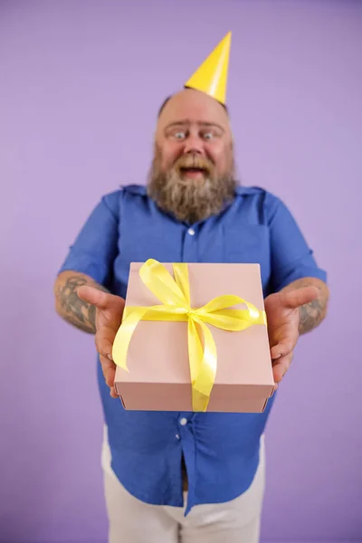 Känslomässig fet man bär hatt står på lila bakgrund, fokus på händer med presentförpackning — Stockfoto
