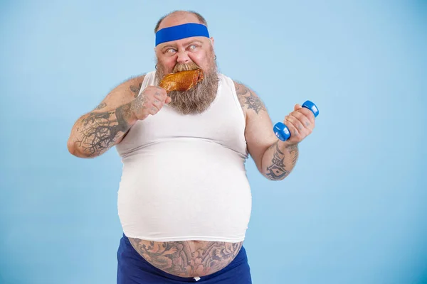 Lustiger Plus Size Mann isst Hühnerbein und macht Übungen mit Hantel auf blauem Hintergrund — Stockfoto