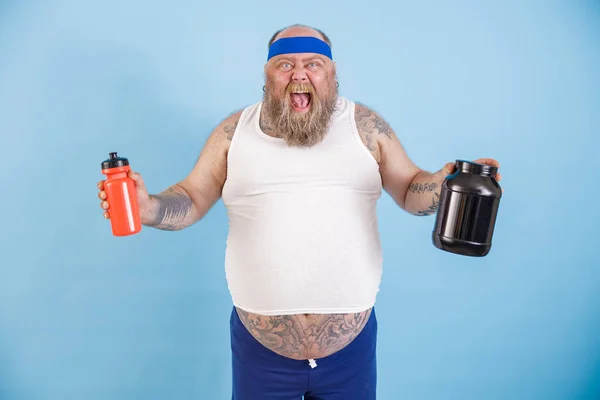 Aufgeregter dicker Mann mit Stirnband hält Getränkeflaschen und Proteinergänzungsmittel im Studio — Stockfoto