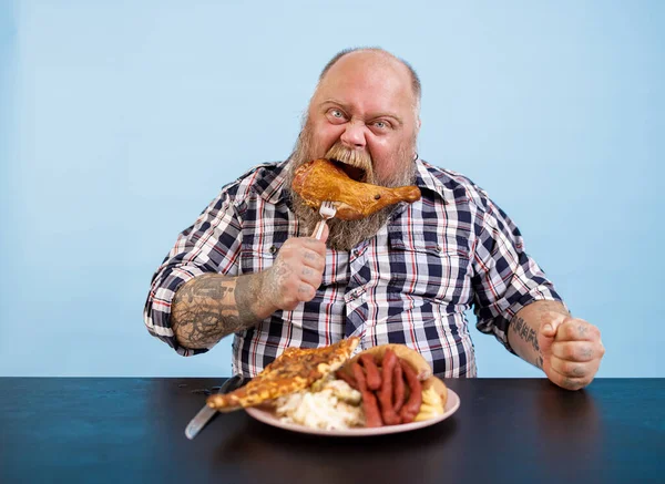 Grappige man met overgewicht eet gerookte kippenpoot aan tafel op lichtblauwe achtergrond — Stockfoto