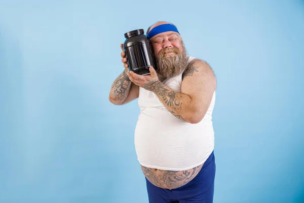 Positiver Mann mit Übergewicht umarmt Flasche Sporternährung auf hellblauem Hintergrund — Stockfoto