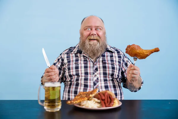 Glimlachende man met overgewicht houdt kippenpoot zittend aan tafel op lichtblauwe achtergrond — Stockfoto