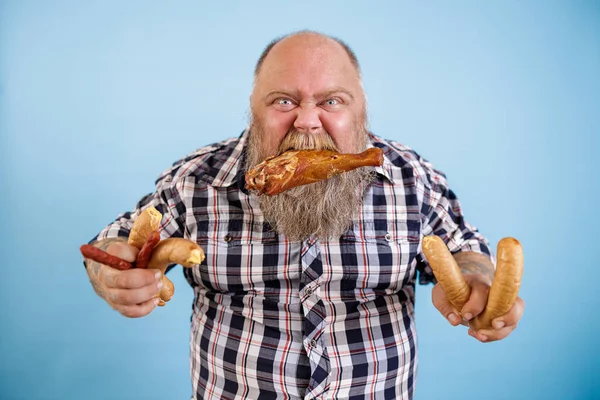 탐욕 스러운 뚱뚱 한 남자는 닭 다리에 이빨 과푸른 배경에 소시지 다발을 들고 있다 — 스톡 사진