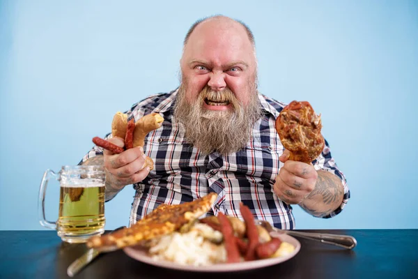 Wütender Mann mit Übergewicht hält Hühnerkeulen und Würstchen mit fettigem Essen am Tisch — Stockfoto