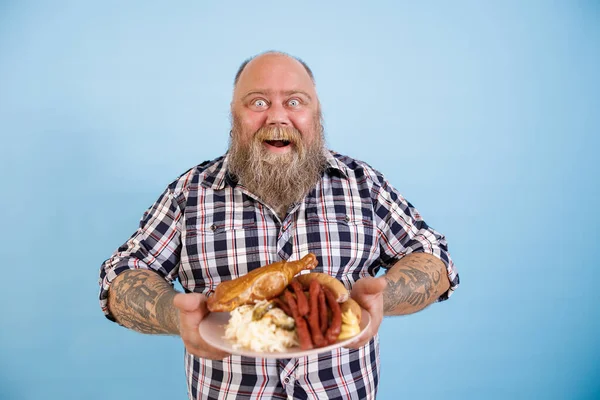 Jouful man met overgewicht houdt plaat van rijk voedsel staande op lichtblauwe achtergrond — Stockfoto