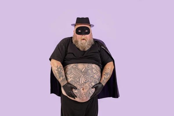 Twijfelende man met overgewicht in carnaval pak houdt enorme buik op paarse achtergrond — Stockfoto