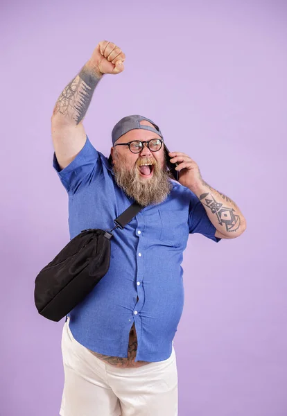 Podekscytowany otyły człowiek z torbą rozmowy na smartfonie podnosząc pięść na fioletowym tle — Zdjęcie stockowe