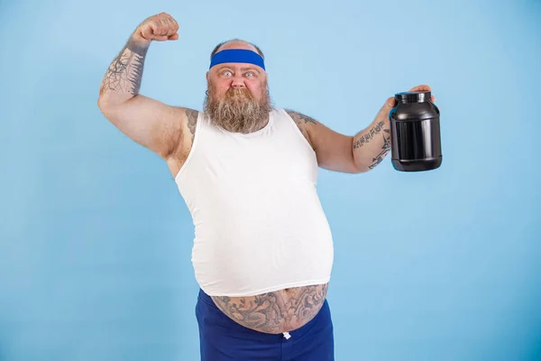 Sterke man met baard met overgewicht toont fles sportvoeding op lichtblauwe achtergrond — Stockfoto