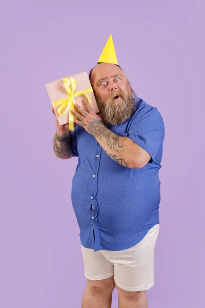 Rolig man med övervikt sätter öra till presentförpackning poserar på lila bakgrund i studio — Stockfoto