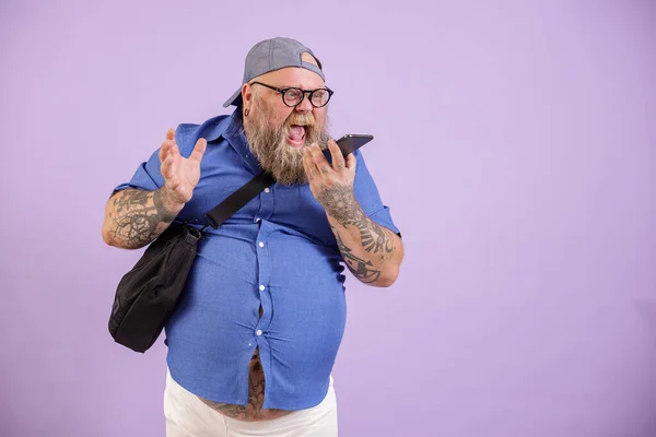 Boze man met overgewicht schreeuwt per telefoon met behulp van luidspreker modus op paarse achtergrond — Stockfoto