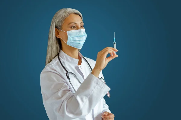 Μεσήλικας γιατρός με ιατρική μάσκα σε μπλε φόντο, επικεντρωθεί σε χέρι με σύριγγα — Φωτογραφία Αρχείου