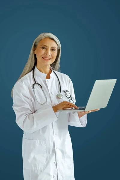 Χαρούμενη ώριμη γιατρός κυρία σε λευκό ρόμπα χρησιμοποιεί σύγχρονο φορητό υπολογιστή στέκεται σε μπλε φόντο — Φωτογραφία Αρχείου