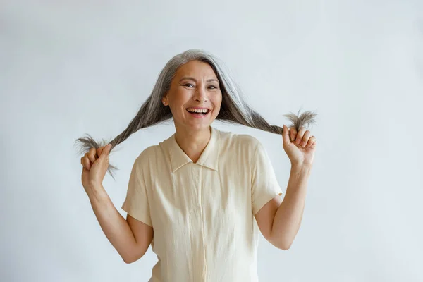 Happy middle aged Asian woman holds twisted hoary hair on light grey background — Stock Photo, Image