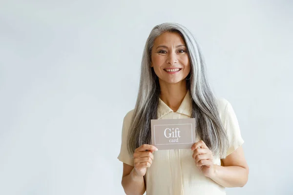 Lächelnde grauhaarige Asiatin hält Geschenkkarte auf hellem Hintergrund — Stockfoto