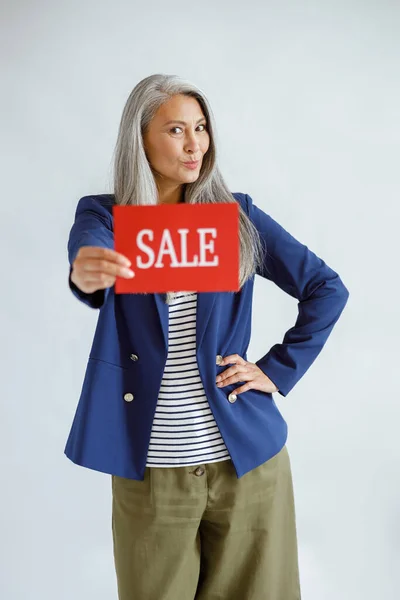 Madura asiática dama con pelo gris en chaqueta sostiene tarjeta roja con palabra Venta en estudio —  Fotos de Stock