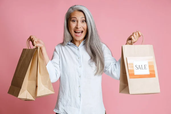 Emocionado mujer asiática madura sostiene bolsas de papel con colorido signo de venta de pie en el estudio — Foto de Stock