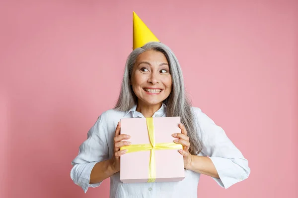 Emocional mujer asiática con pelo gris suelto y sombrero de fiesta sostiene presente sobre fondo rosa — Foto de Stock