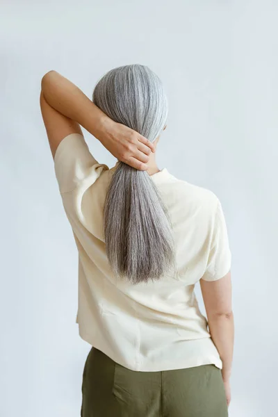 Woman in yellow shirt makes polytail of silver hair standing on light grey background — Stock Photo, Image