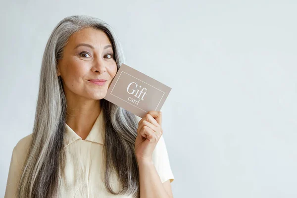 喜庆的灰色头发的亚洲女人拿着礼品卡，摆出淡灰色背景 — 图库照片