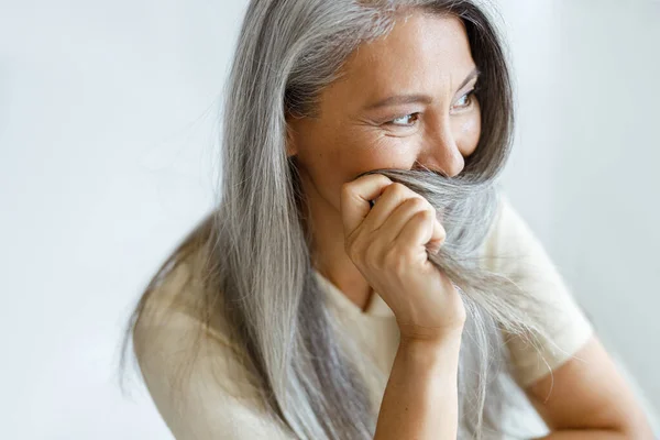 Middelbare leeftijd aziatische vrouw verbergt gezicht met grijs haar zitten op lichte achtergrond — Stockfoto