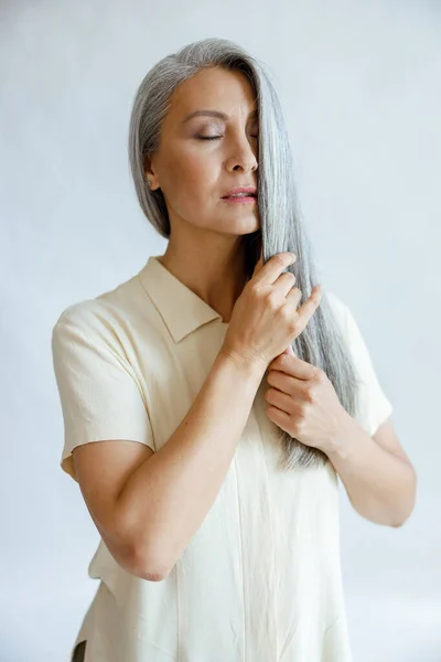 Languid medelålders asiatisk kvinna stroke lås av silver hår på ljusgrå bakgrund — Stockfoto