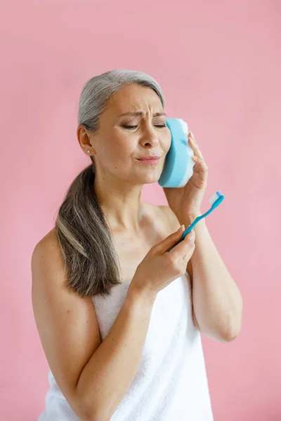 Walgelijke middelbare leeftijd aziatische vrouw kijkt naar tandenborstel holding bad spons nera oor in studio — Stockfoto