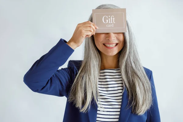 满脸笑容，头发蓬松的成熟女士摆着淡灰色背景的礼品卡 — 图库照片