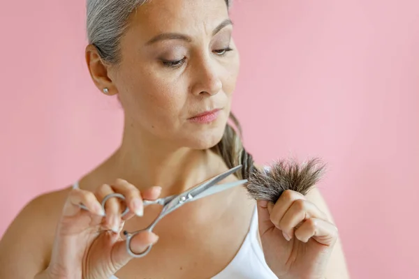 Zweifelnde Asiatin schneidet ausgefranste Enden langer grauer Haare auf rosa Hintergrund im Studio — Stockfoto