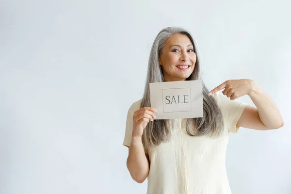 Alegre mujer asiática de mediana edad con puntos de pelo gris en venta signo sobre fondo claro —  Fotos de Stock