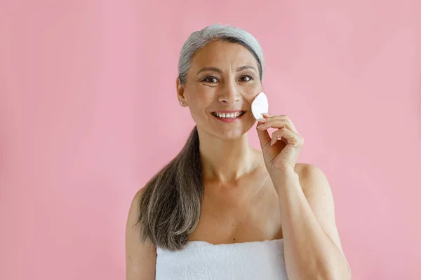 Sorrindo de cabelos grisalhos mulher asiática limpa o rosto com almofada de algodão em pé sobre fundo rosa — Fotografia de Stock