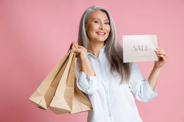 Hermosa señora de mediana edad sostiene bolsas de papel y tarjeta con palabra Venta en estudio —  Fotos de Stock