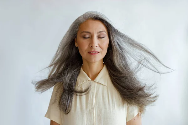 Pretty tranquil Asian woman with flying silver hair stands on light grey background — Stock Photo, Image