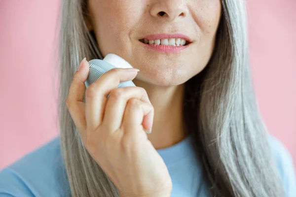 En lykkelig middelaldrende kvinne vasker ansiktet med en sonisk pensel på rosa bakgrunn – stockfoto