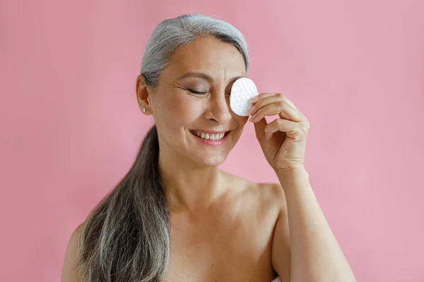 Positieve grijze haren Aziatische dame verwijdert make-up uit het oog met katoen pad in de studio — Stockfoto