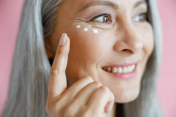 Schöne reife asiatische Dame trägt Anti-Aging-Creme unter den Augen auf rosa Hintergrund auf — Stockfoto